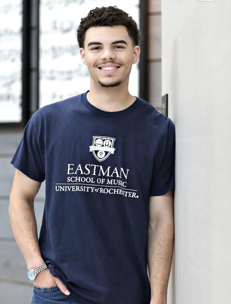 A man in a navy blue t-shirt smiles and leans against a wall. The shirt has white text.