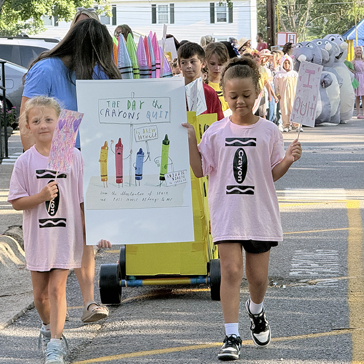 Two children in pink shirts walk in a parade with a cart displaying a colorful drawing.