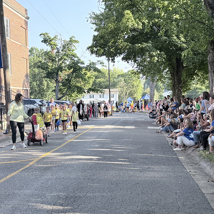 A group of people in yellow shirts walk down a street. Spectators sit and stand on the side.