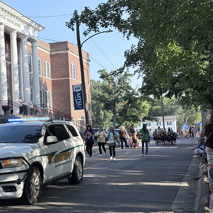 A sheriff's SUV is parked on a street, with a crowd of people walking nearby.