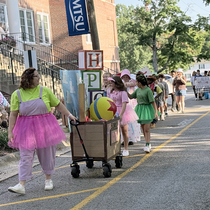 People in costumes and tutus walk in a parade, pushing a cart with a ball.