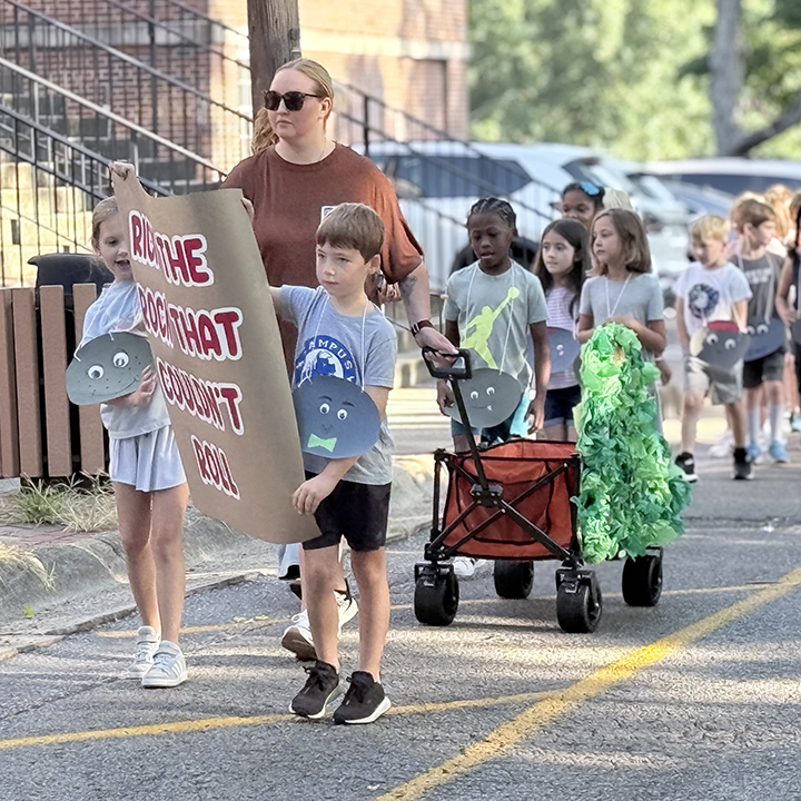 A group of children and adults walking on a street. Some carry a sign, and others push carts.