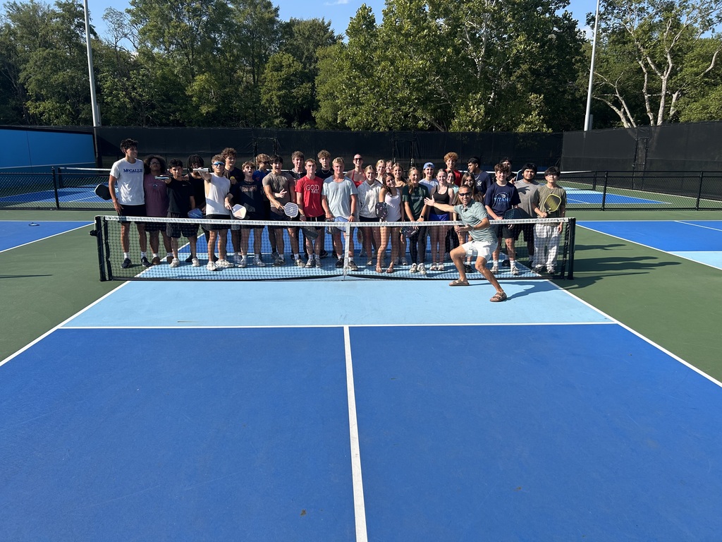 A group of people stands on a tennis court with blue and green surfaces. One person is positioned on the net.