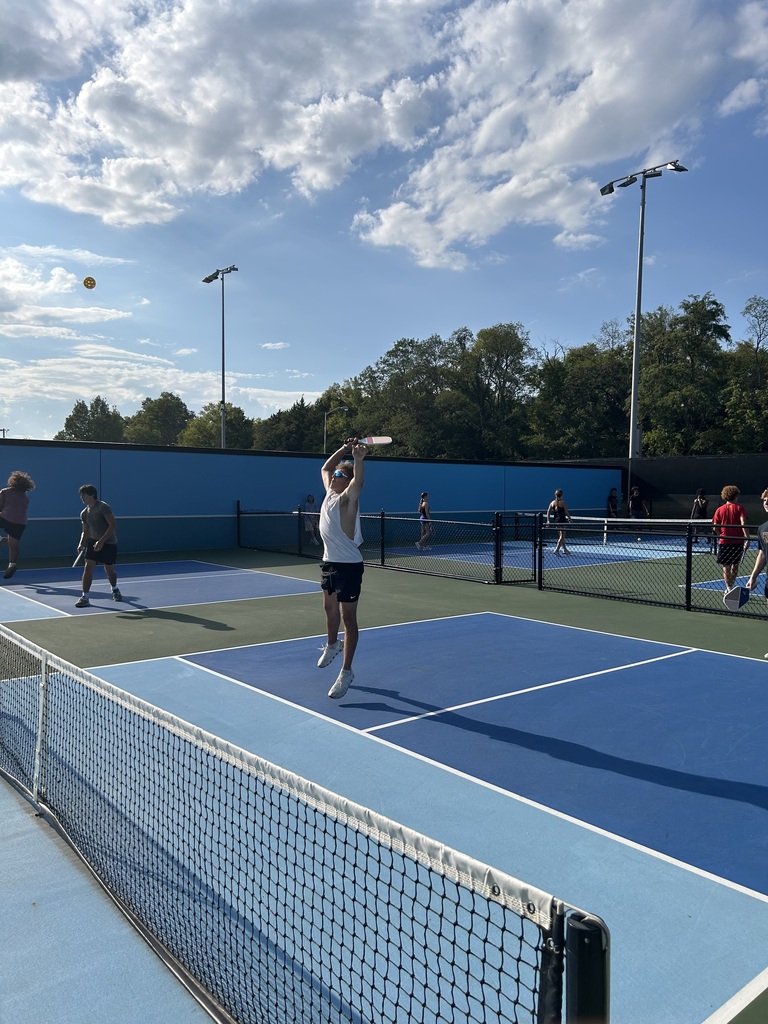 Multiple people playing tennis on an outdoor court with blue and white markings. A player leaps to hit a ball.