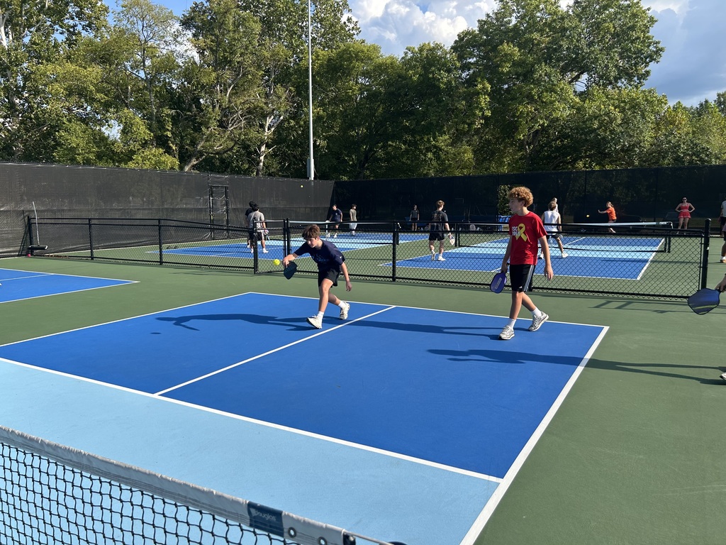 Multiple individuals playing tennis on blue courts, with trees in the background and a fence surrounding the area.