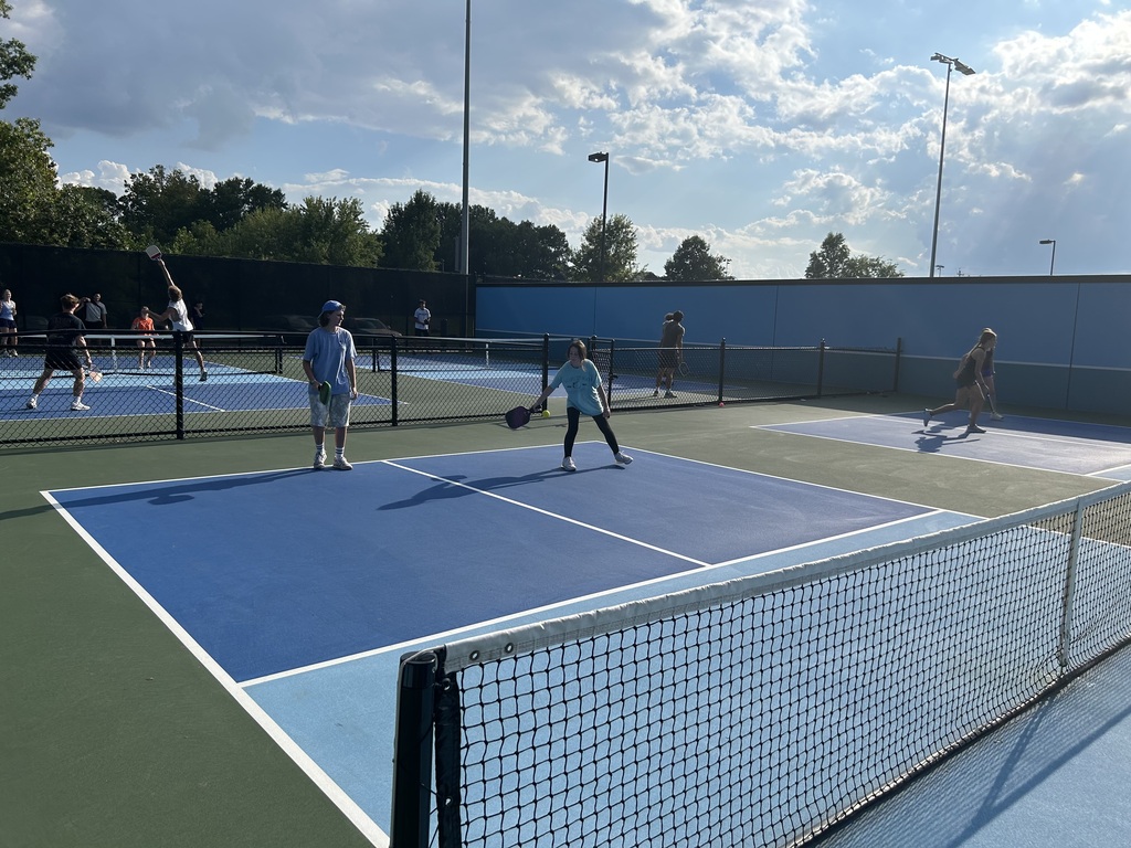 An outdoor tennis court with several people playing tennis. There are blue and white markings on the court and a net in the middle.