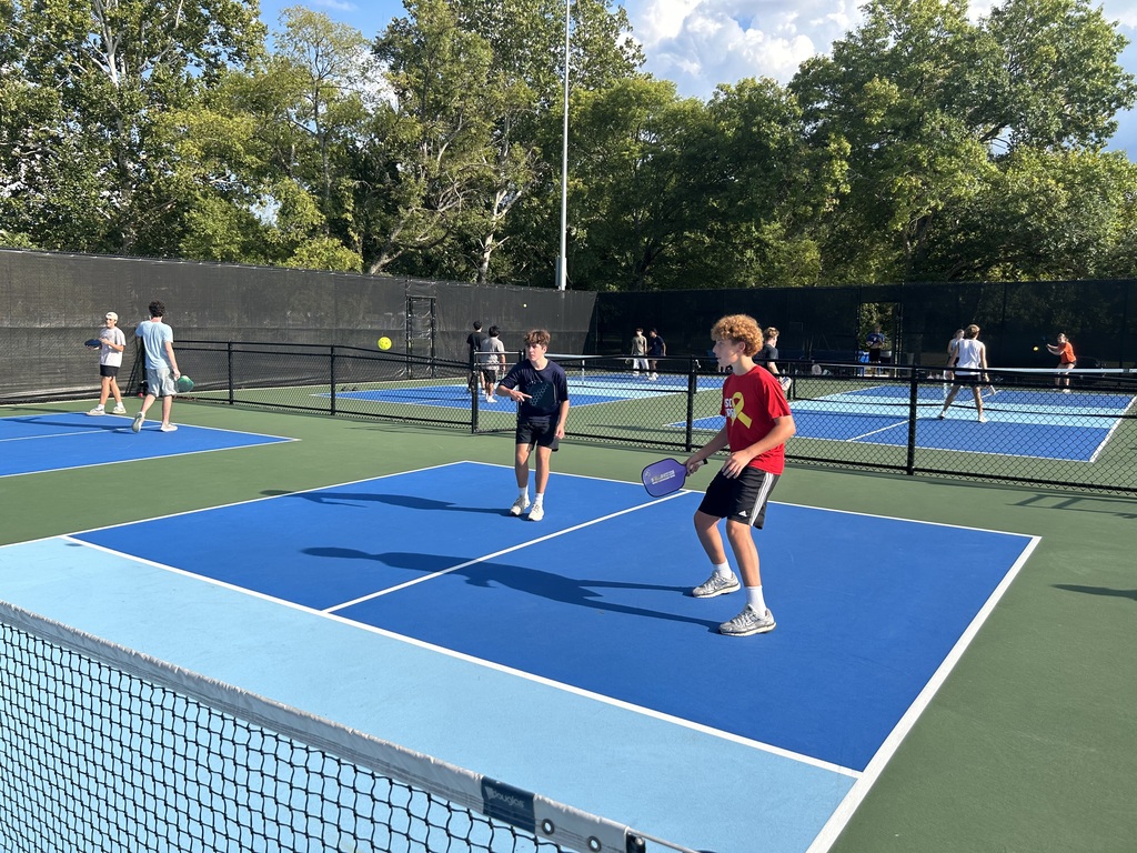 Multiple tennis players on blue and green courts. One player in red holds a racket, preparing to hit the ball.