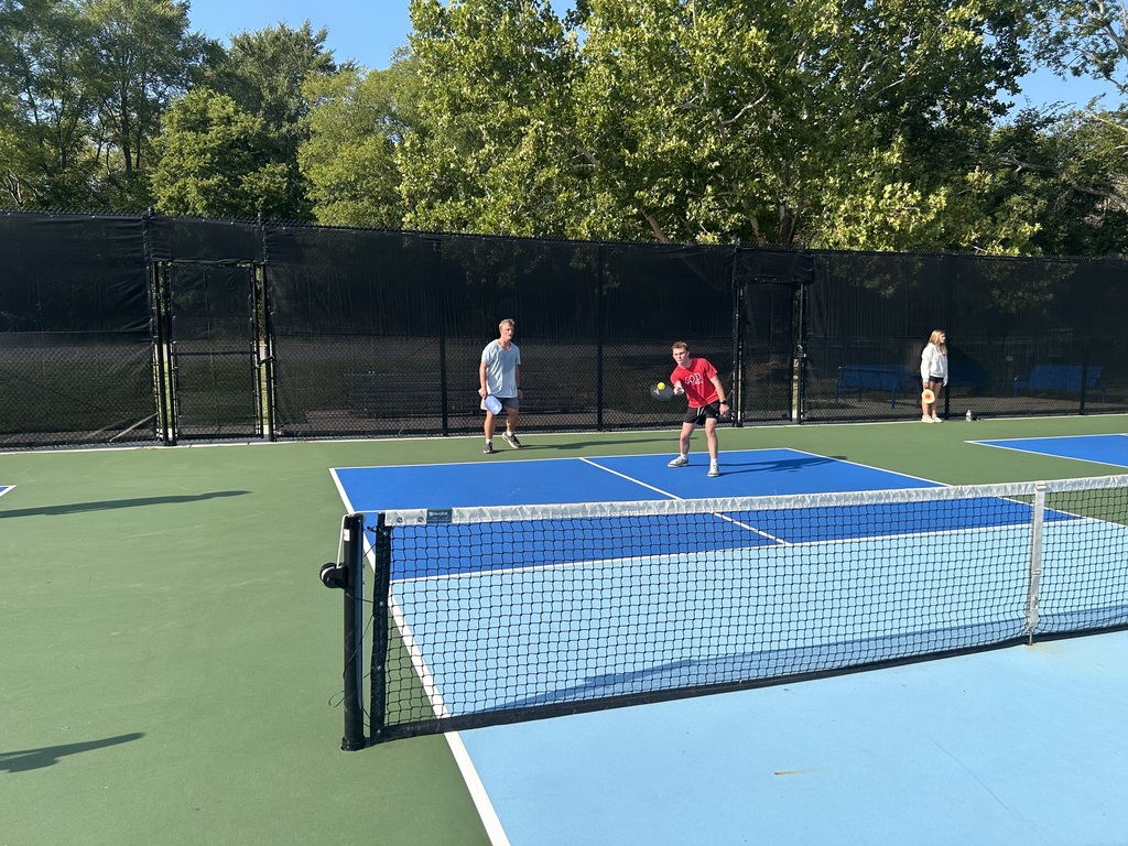 Two players in red and white gear play on a blue and white outdoor tennis court. Two others stand on the sidelines.