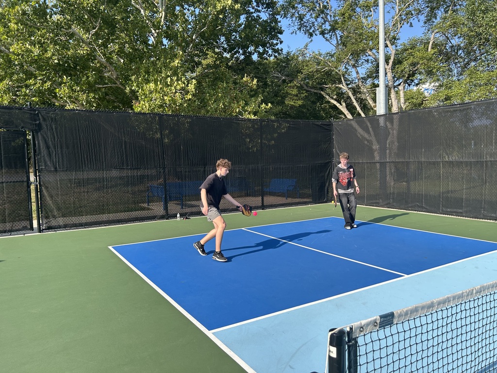 Two people play table tennis on a blue court with a fence and trees in the background.