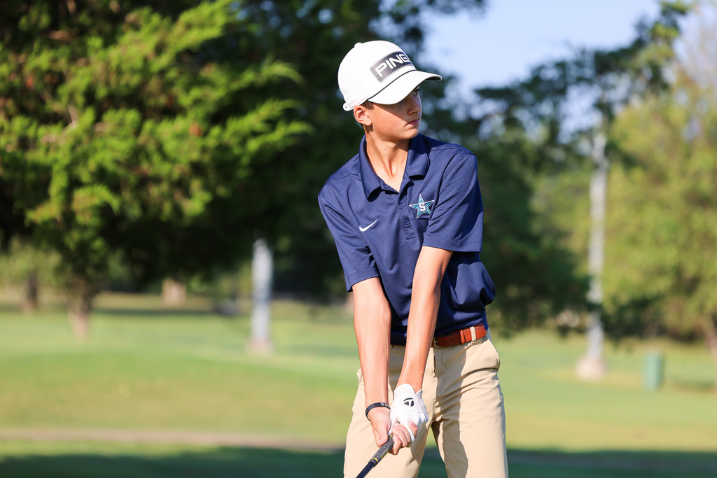 Person in blue shirt, khaki pants, and a white cap swings a golf club on a golf course.
