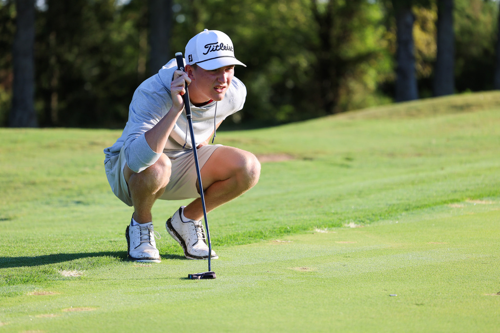 Person wearing a hat, long-sleeved shirt, shorts, and white shoes, crouching to inspect a golf ball.