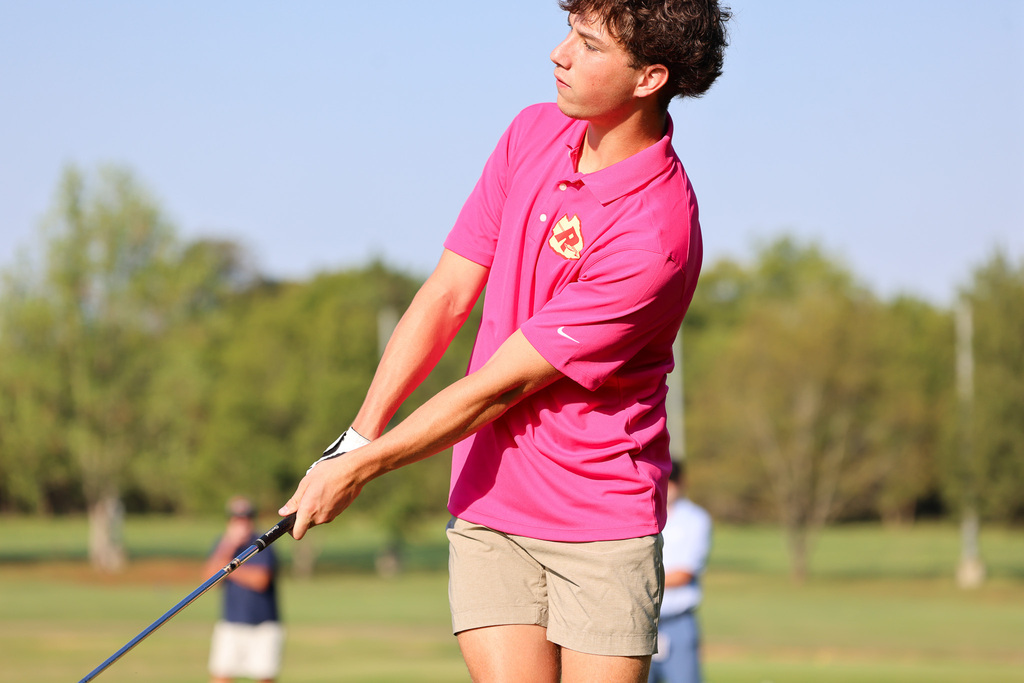 A man swings a golf club in a field with trees in the background. Two people are blurred in the background.