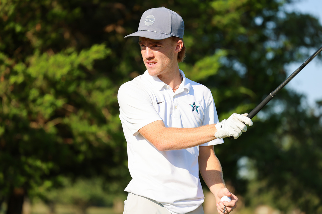 A man in a white polo with a star on the left chest, a gray cap, and white gloves holds a golf club.