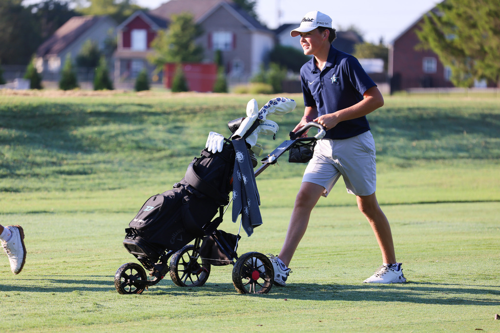 A person in blue shirt and white shorts pushing a golf trolley on a golf course.