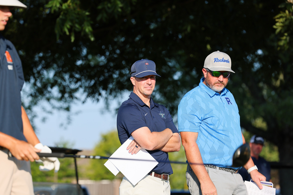 Three men in hats and belts stand by a fence; one holds papers, another holds a golf club.