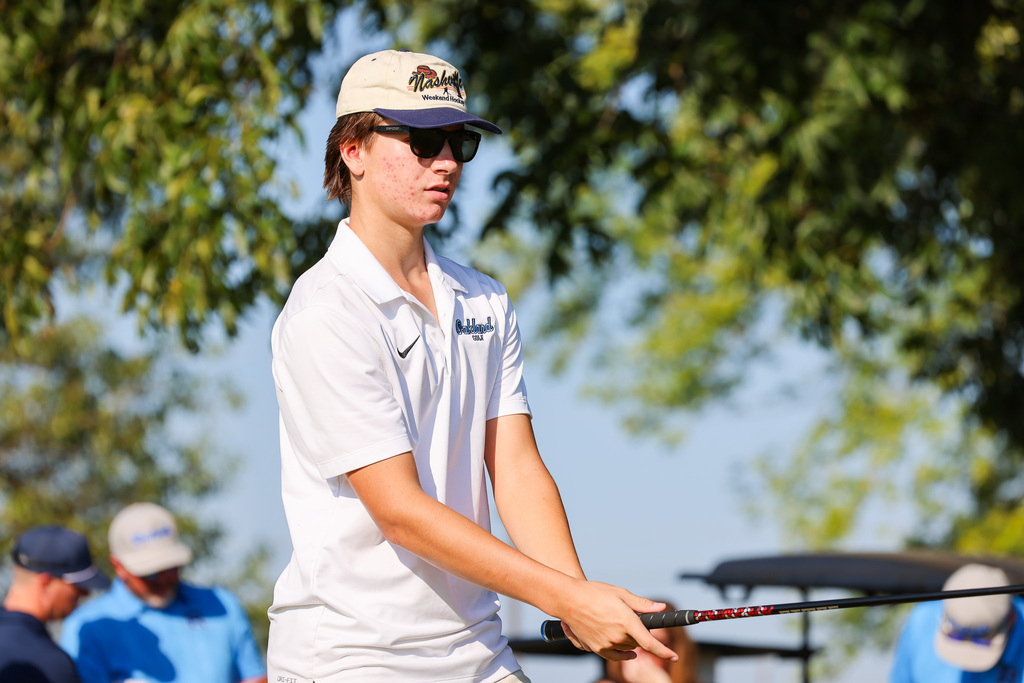 Person in a white polo shirt and cap, holding a golf club. Background includes trees and people in blue shirts.