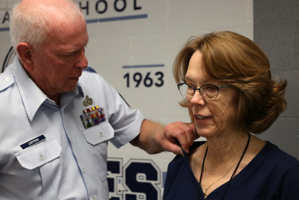 A man in a white uniform pins a medal on a woman wearing glasses. The background displays text and numbers.