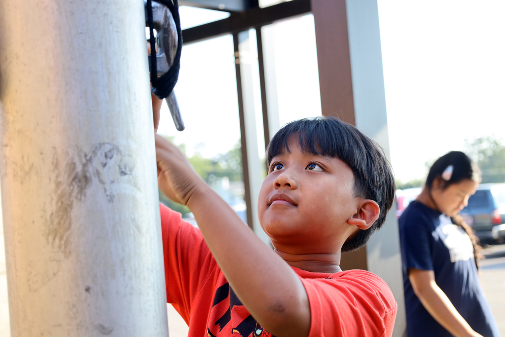 A boy in an orange shirt climbs a metal pole, looking up. A girl in a blue shirt stands nearby.