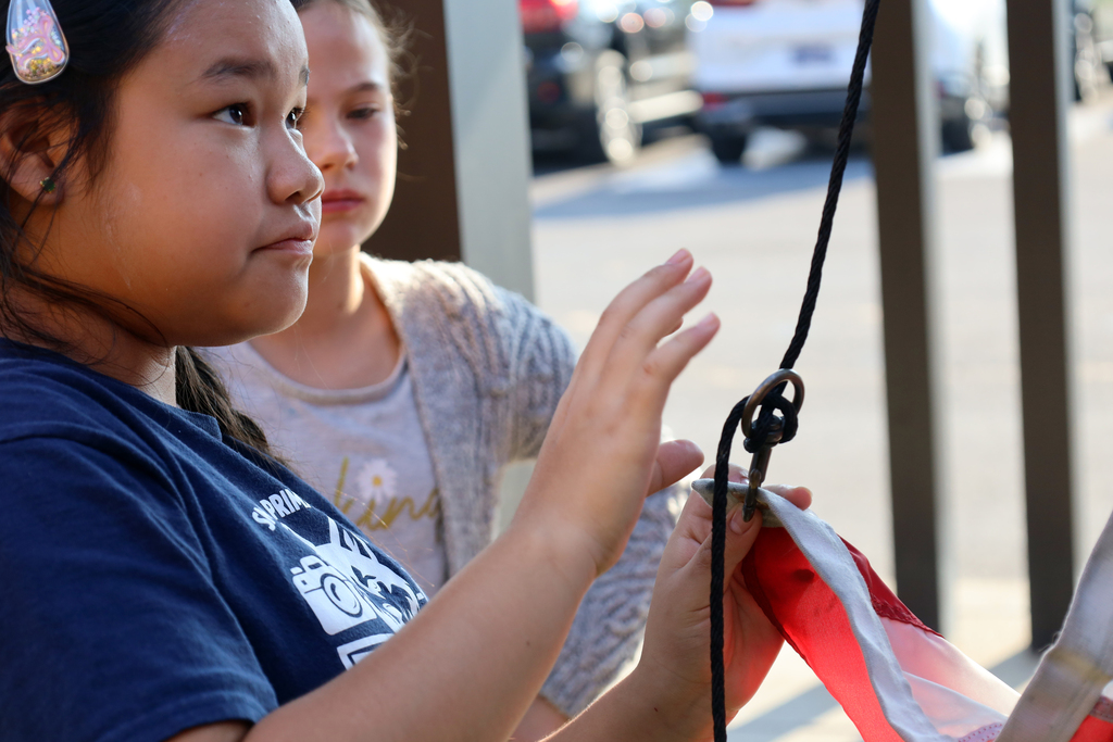 Two children, one holding a rope swing, stand outdoors in a parking lot with cars visible in the background.