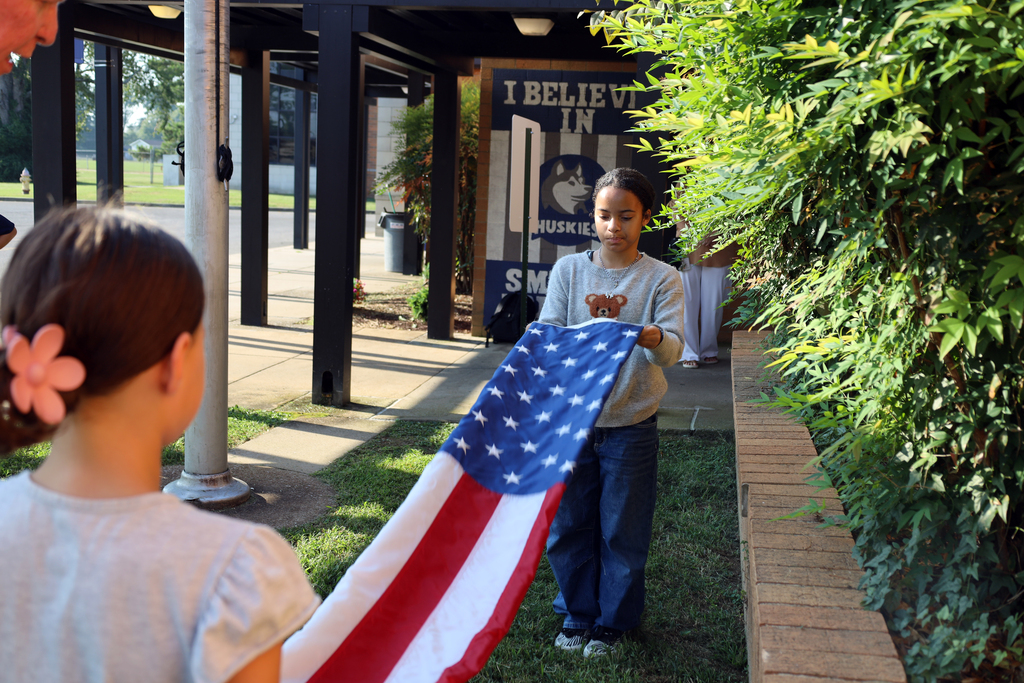 Two girls hold an American flag in a schoolyard. A sign reading "I BELIEVE IN DOUGLAS" is behind them.
