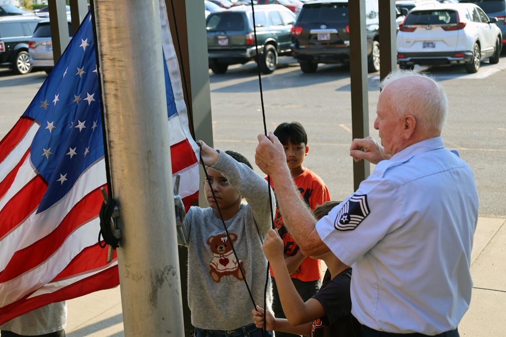 A man assists two children in raising a flag on a pole, with cars parked in a lot in the background.