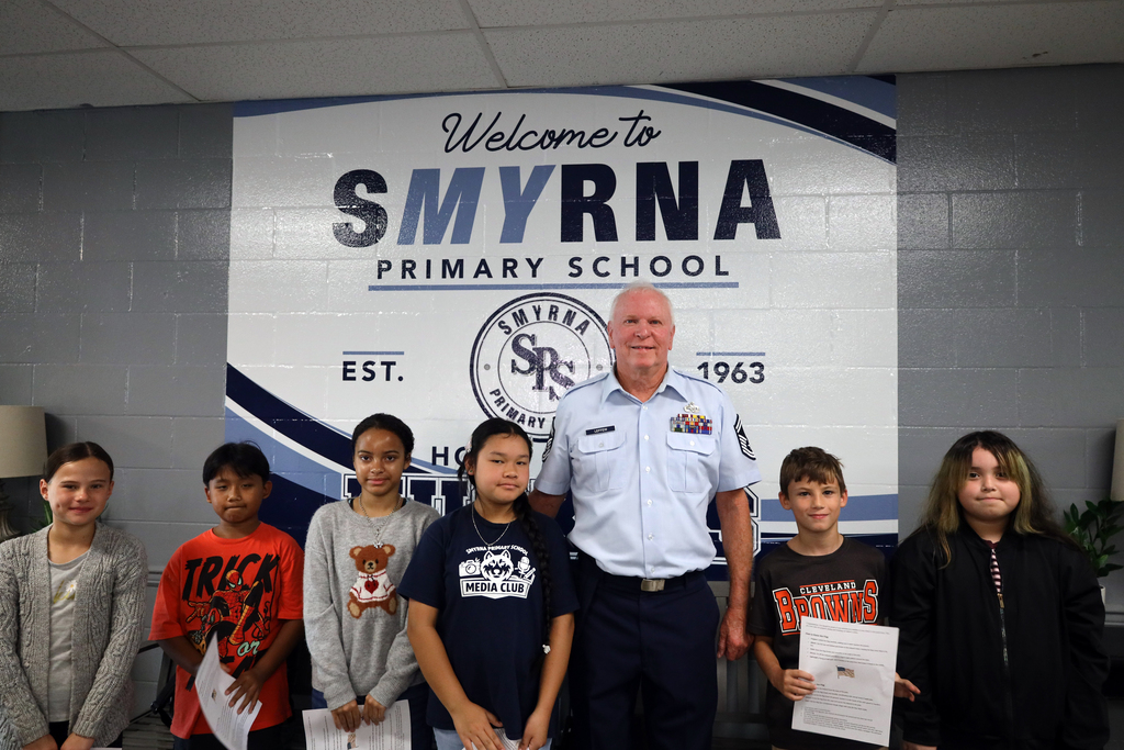 A man in uniform stands with six children in front of a sign for Smyrna Primary School.