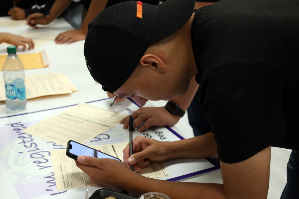 A man in black, wearing a cap, writes on a paper with a pen. A phone rests on the paper.