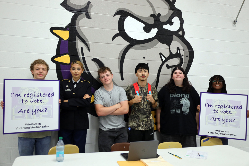 Five people hold signs saying "I'm registered to vote. Are you?" near a large school mascot mural.