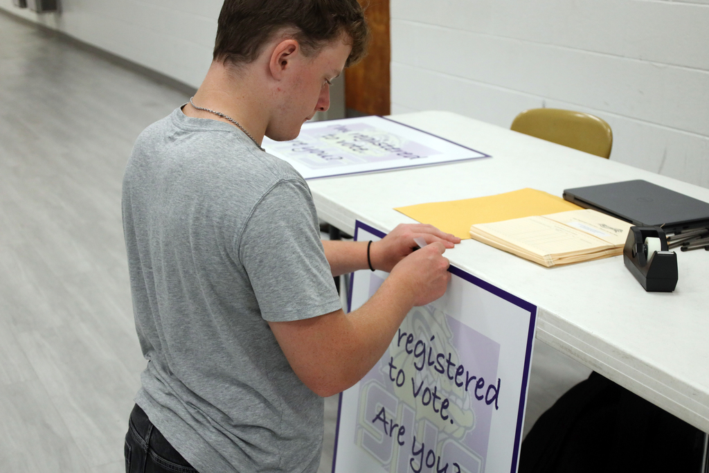 A person in a gray shirt adjusts a sign that reads "Registered to vote. Are you?" in a room.