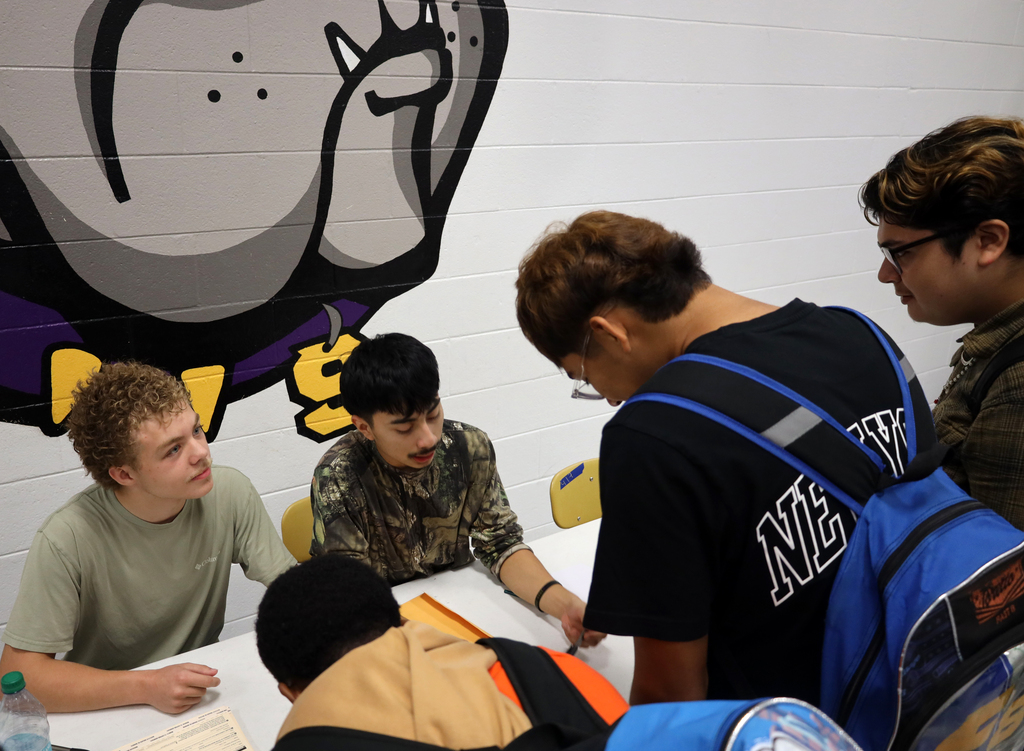 Five individuals are gathered around a table with papers and water bottles. The wall behind them features a large, cartoonish drawing.
