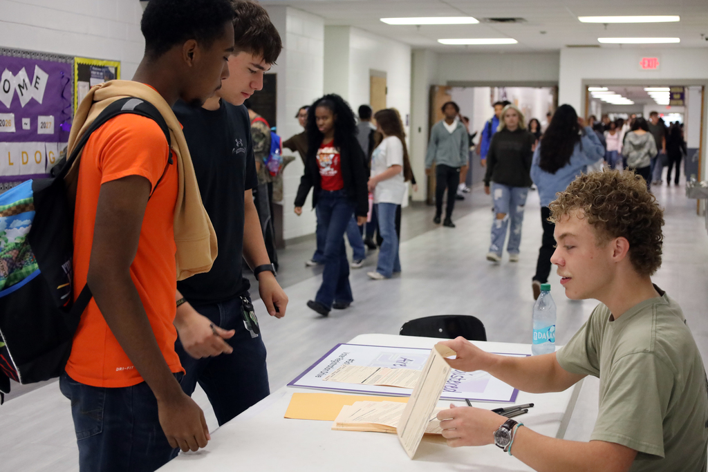Students walking in a school hallway, one sits at a desk while another stands behind him.