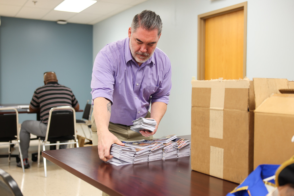 A man in a purple shirt organizes stacks of pamphlets on a table, with chairs and boxes nearby.
