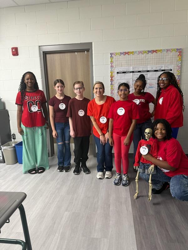 Nine children and one adult pose in a classroom with a bulletin board and door. Some hold skeletons.