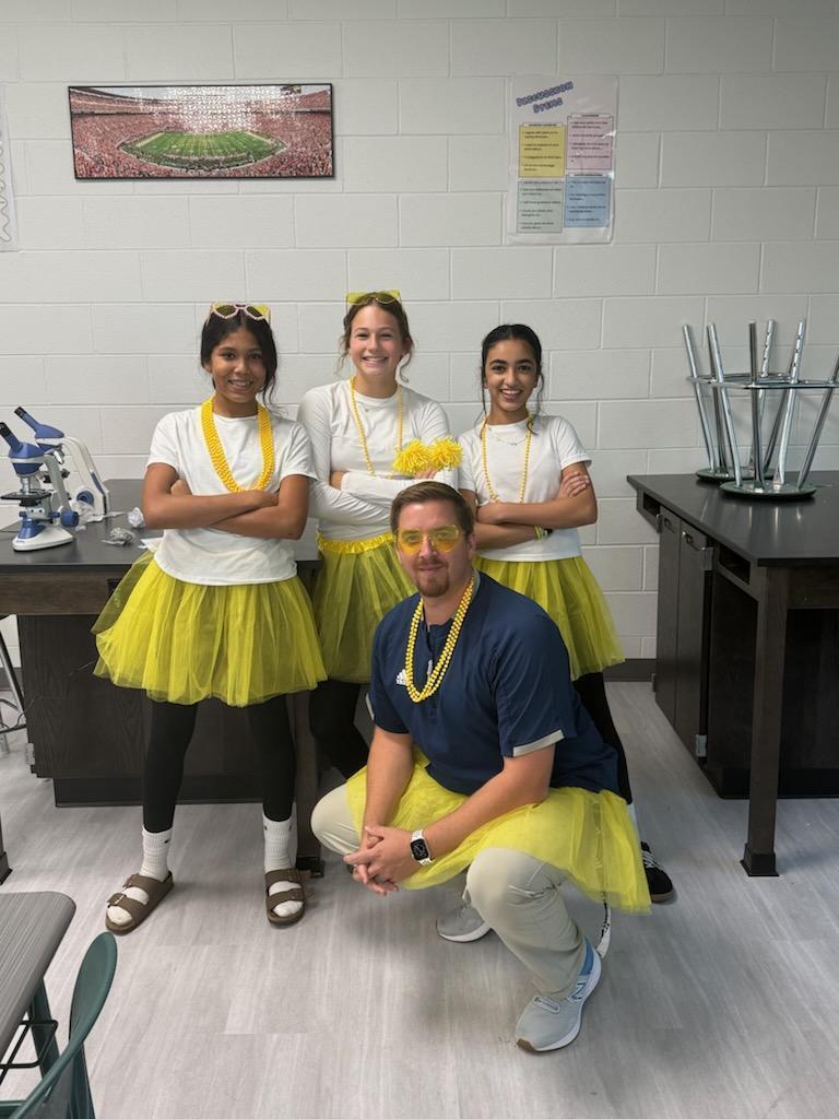 Four people in a classroom, three wearing matching yellow tutus, posing with a man in a blue shirt.