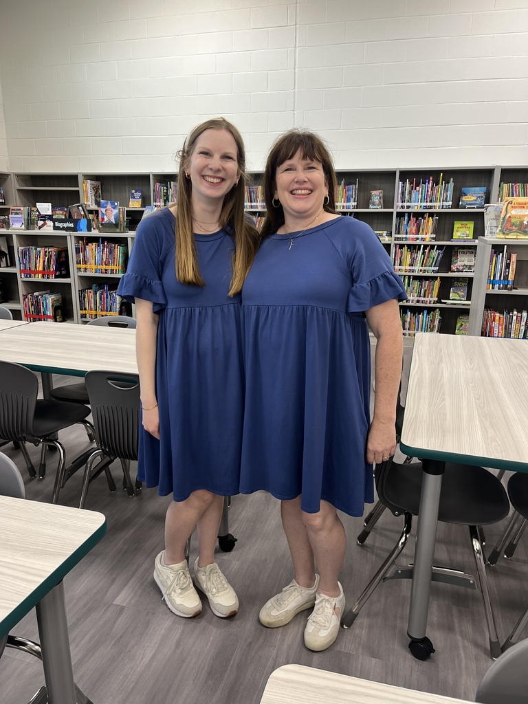 Two women in blue dresses and white shoes stand in a library, surrounded by tables and chairs.