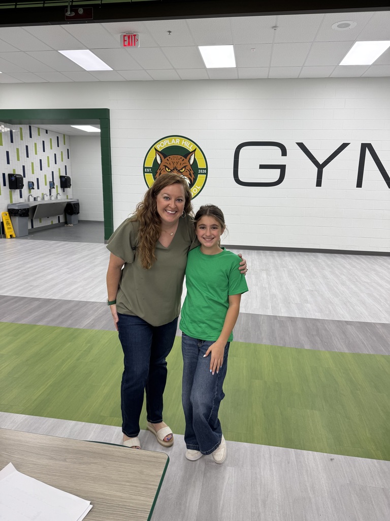 Two people stand in a gym. One wears green, the other blue. Behind them, a wall displays a large "GYM" sign with a logo.