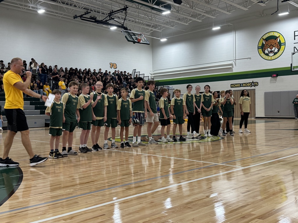 A group of children in green uniforms on a basketball court. A man in yellow stands with a microphone.