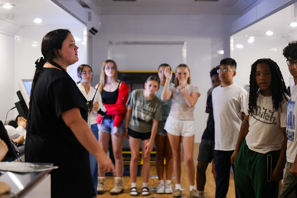 A woman in black speaks to a group of students in a room with white walls and wooden floors.