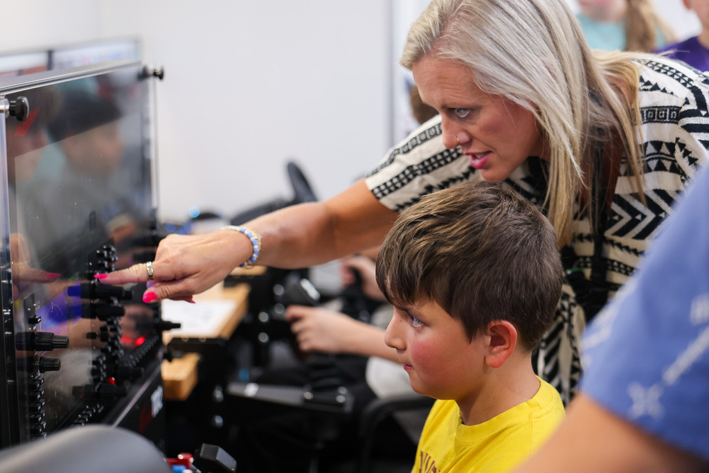 Woman with blond hair points at monitor with a boy in a yellow shirt. Background shows blurred people.