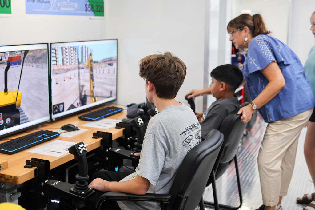 A woman stands behind two kids using a computer in a lab. The screen shows a simulated construction site.