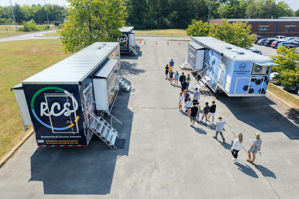 People walk towards mobile units parked in a parking lot. Two trucks have large, colorful graphics. A building and parked cars are in the background.