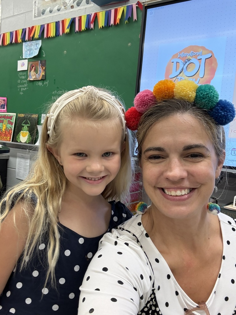 A girl and a woman pose for a picture, smiling. The girl wears a headband. Behind them is a classroom with a chalkboard and screen.