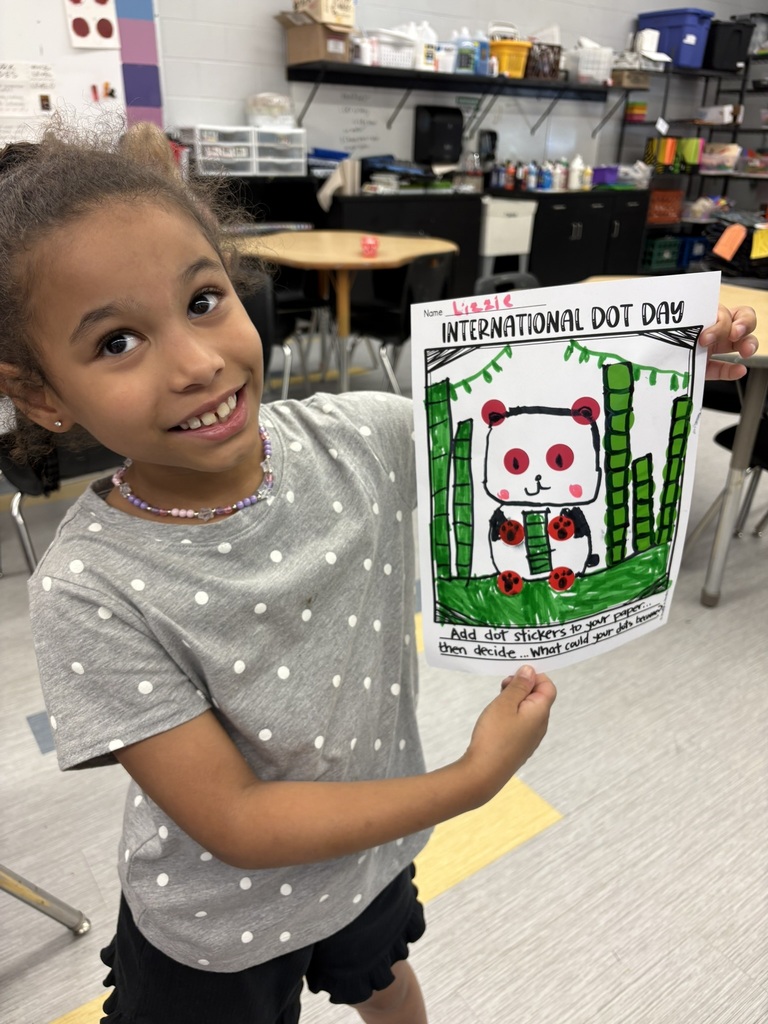 Child in classroom holds up a paper drawing of a panda in bamboo with the words International Dot Day.