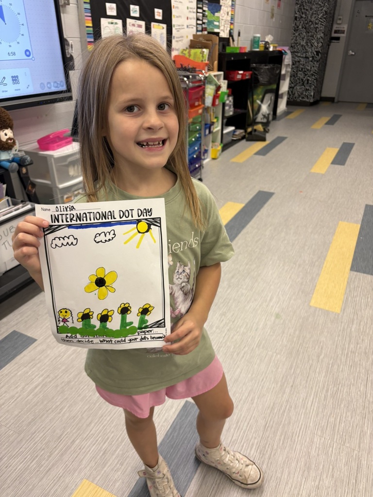 A young girl smiles as she holds a colorful drawing of flowers. The background shows a classroom setting.