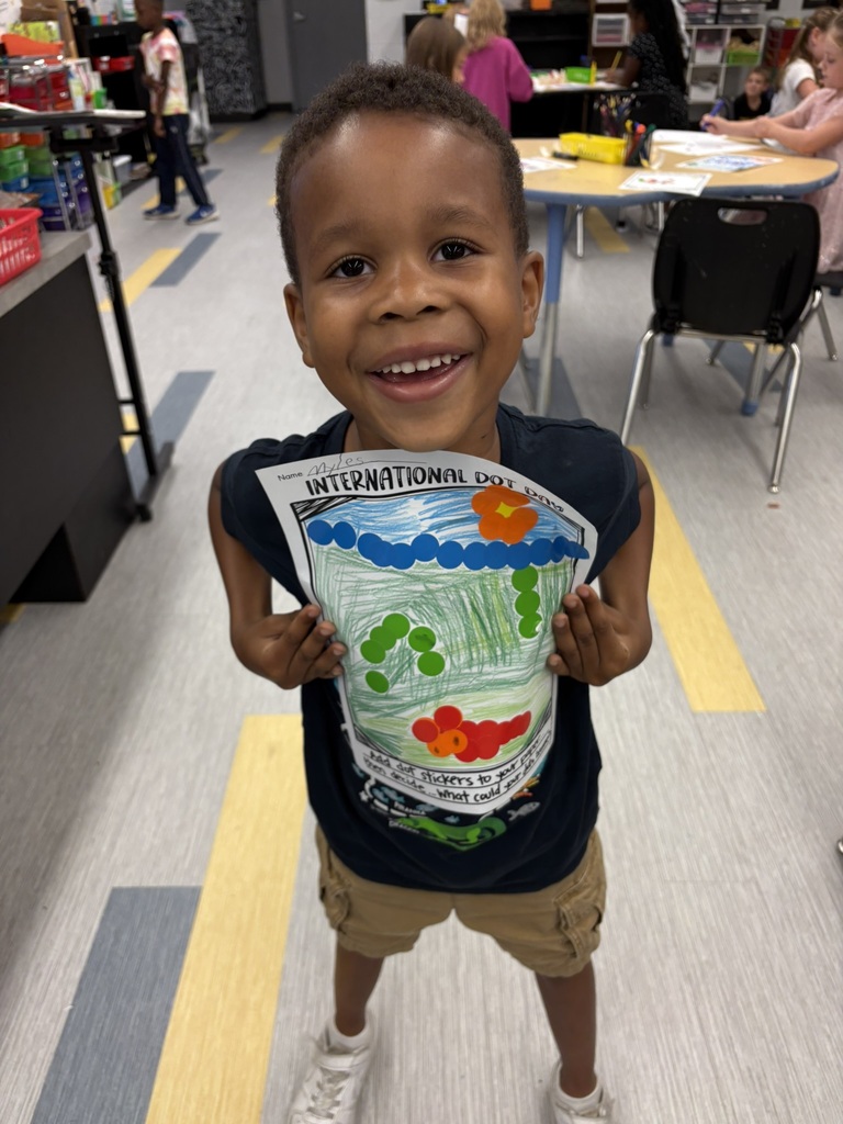 A young boy with a smile holds up a piece of paper with drawings. Behind him, tables and chairs are visible.