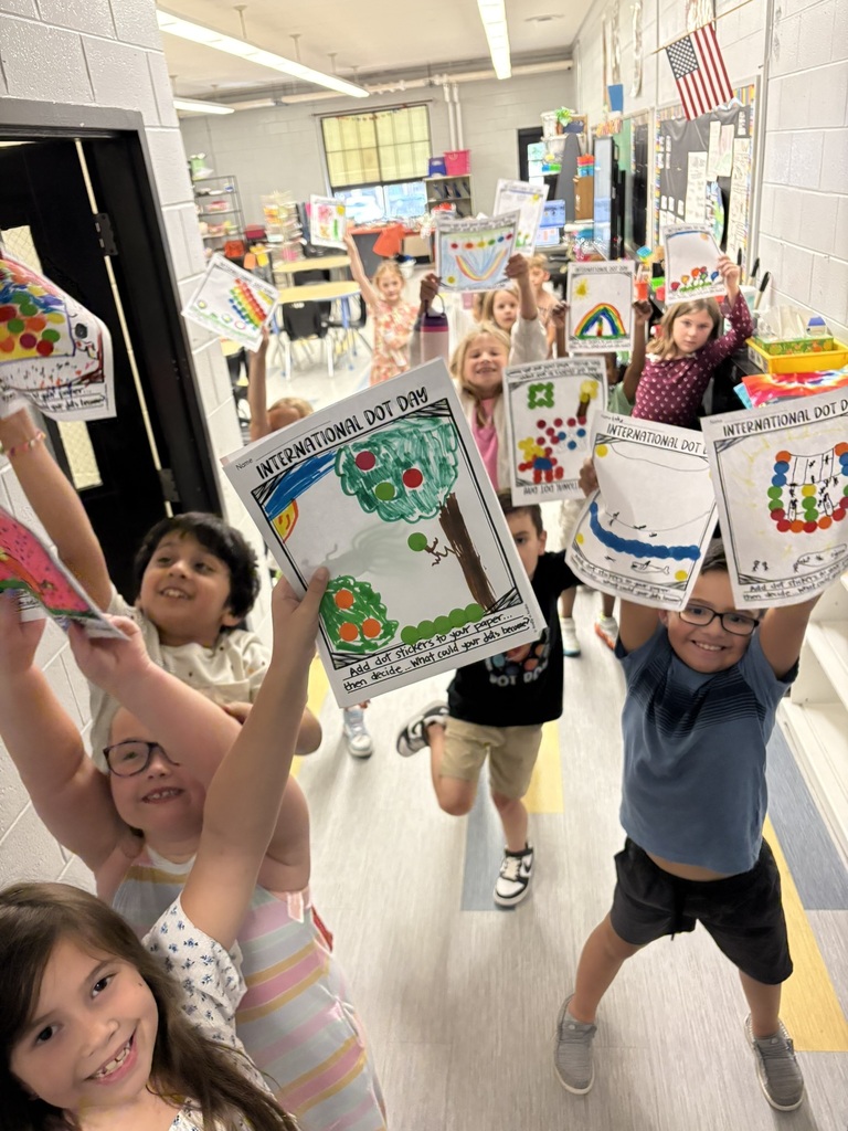 Kids hold up drawings of apples in a hallway. Many are wearing glasses. A flag hangs on the wall.