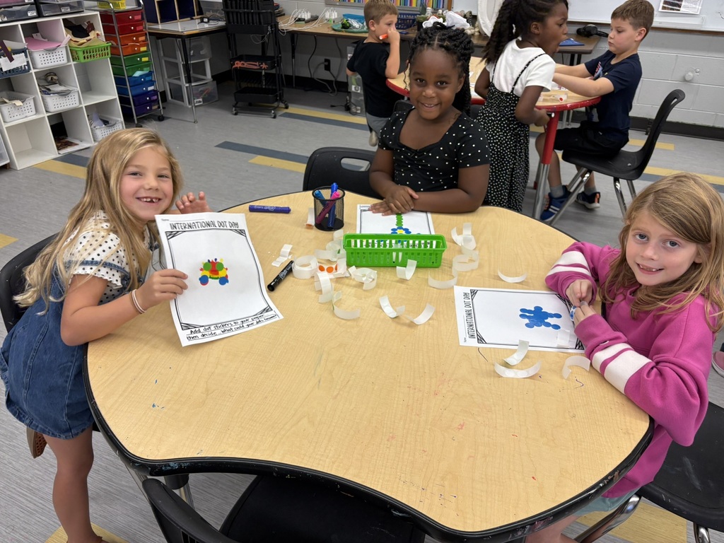 Three girls seated at a table, engaged in a craft activity with cut paper and markers.