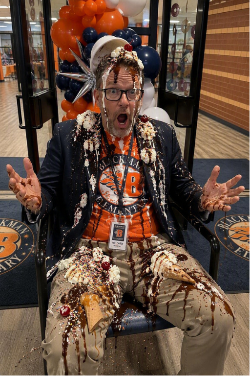 Man in orange shirt, blazer, and khakis sits in chair with ice cream on his body.