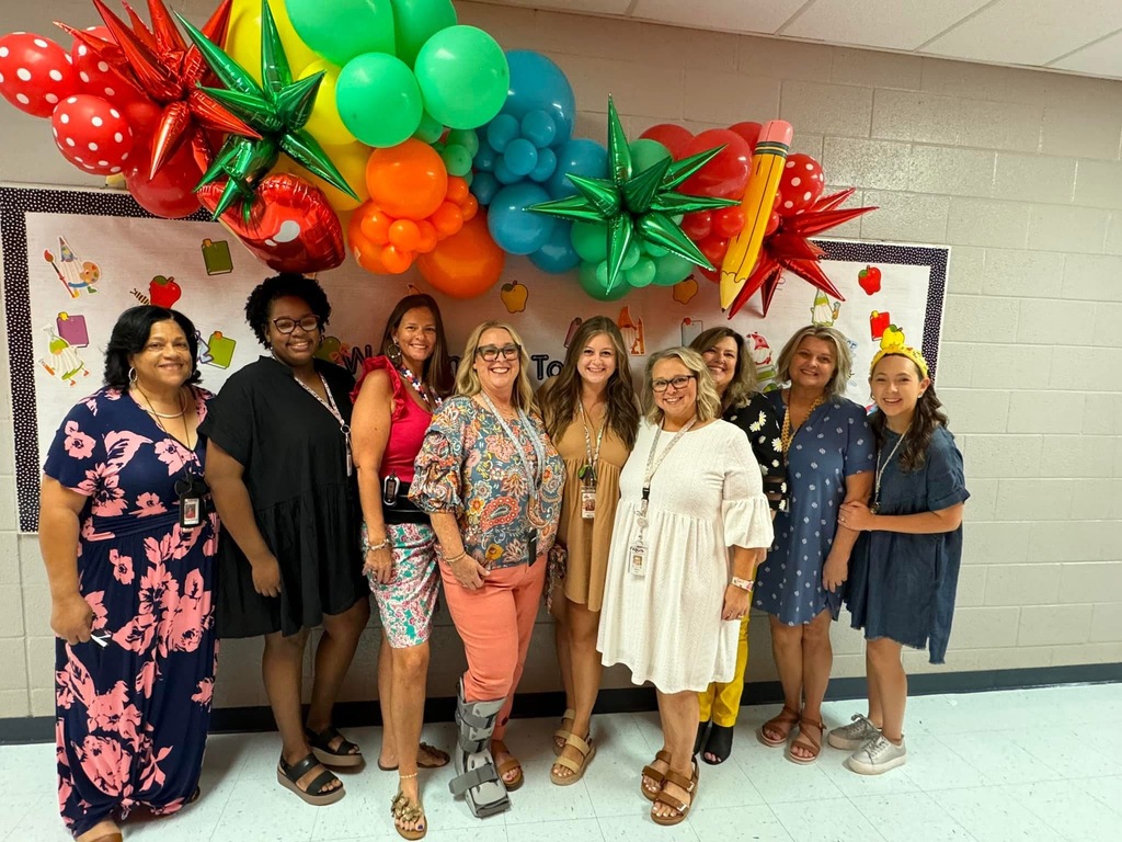 A group of women pose together in front of balloons. They wear casual summer clothing.