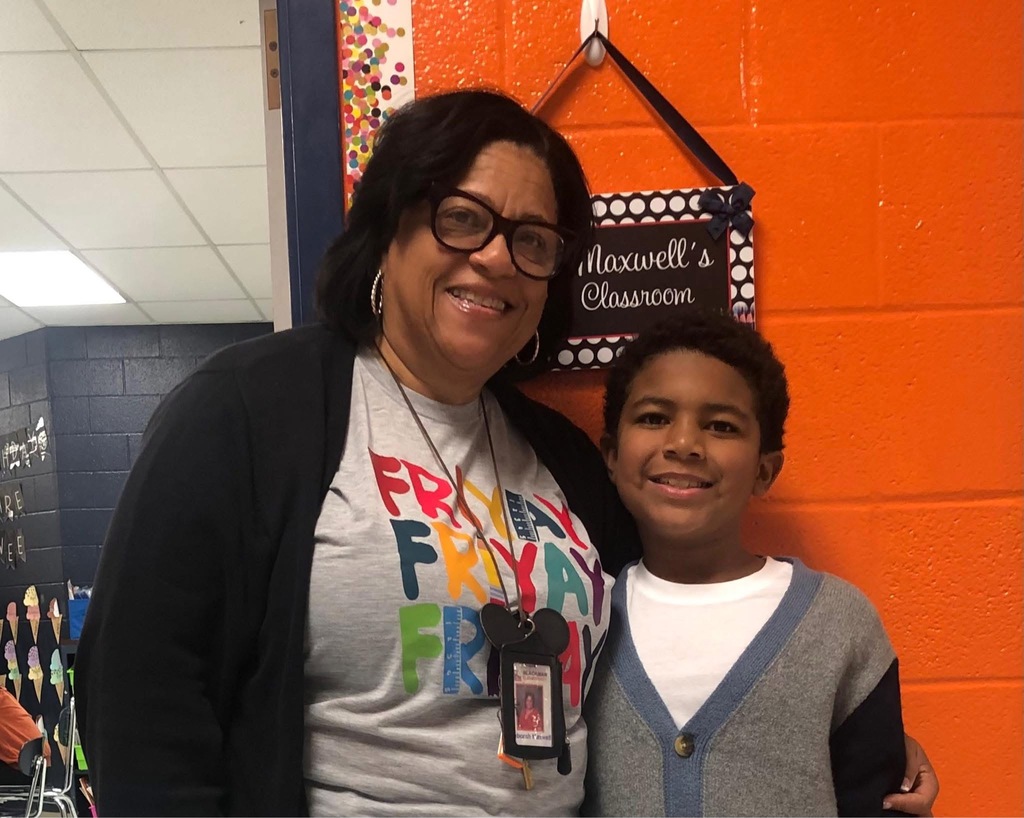 A woman wearing glasses and a gray shirt poses with a boy in a gray sweater. They stand next to an orange wall with a classroom sign.